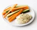 A round, white plate is covered in ingredients for a snack, including a vegan hemp hummus and raw crudites.