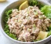 A speckled white bowl holding fresh salmon salad that is laying on top of healthy green leafy lettuce with a couple of lemon wedges.