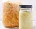 Two jars of homemade sauerkraut sitting on marble surface with white background with one jar filled with carrot along with sauerkraut and the other smaller jar plain.