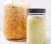 Two jars of homemade sauerkraut sitting on marble surface with white background with one jar filled with carrot along with sauerkraut and the other smaller jar plain.